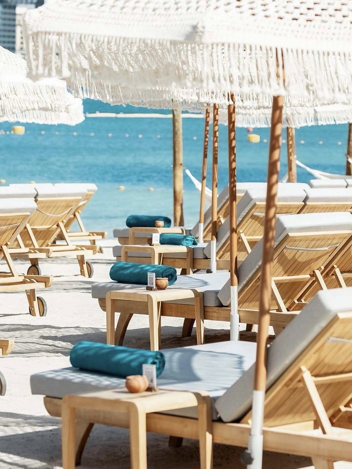 Luxury beach club with wooden loungers, white umbrellas, blue towels on tables, and turquoise sea view