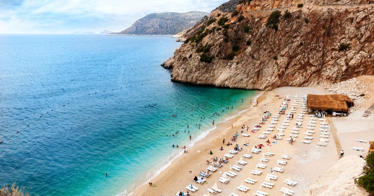 Aerial view of a crowded beach nestled between tall cliffs with turquoise water and organized sunbeds