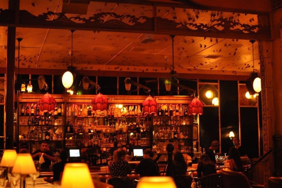 Warm-lit bar interior with illuminated shelves of bottles, hanging red decorations, customers at counter, and ambient yellow lighting in evening setting