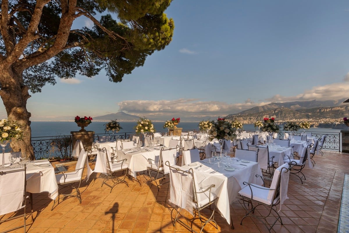 Elegant outdoor wedding reception setup on a terrace overlooking a Mediterranean coastal town with mountains and sea in the background