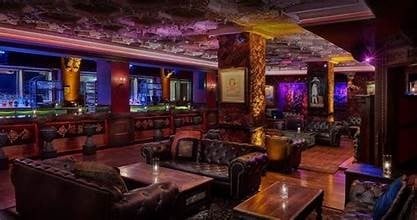 Upscale nightclub interior with purple and gold lighting, leather seating, ornate columns, and city views through windows