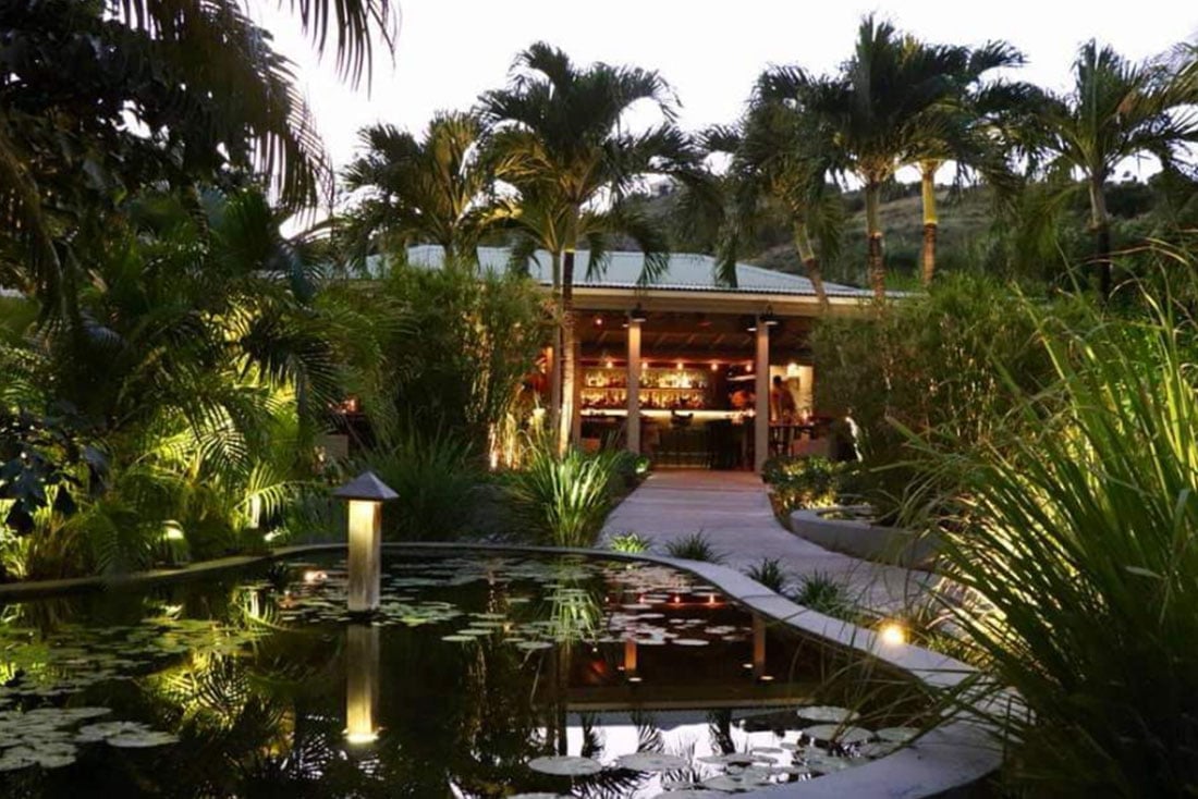 Tropical garden pavilion at dusk with illuminated structure surrounded by palm trees, lush plants, and a reflective pond in the foreground