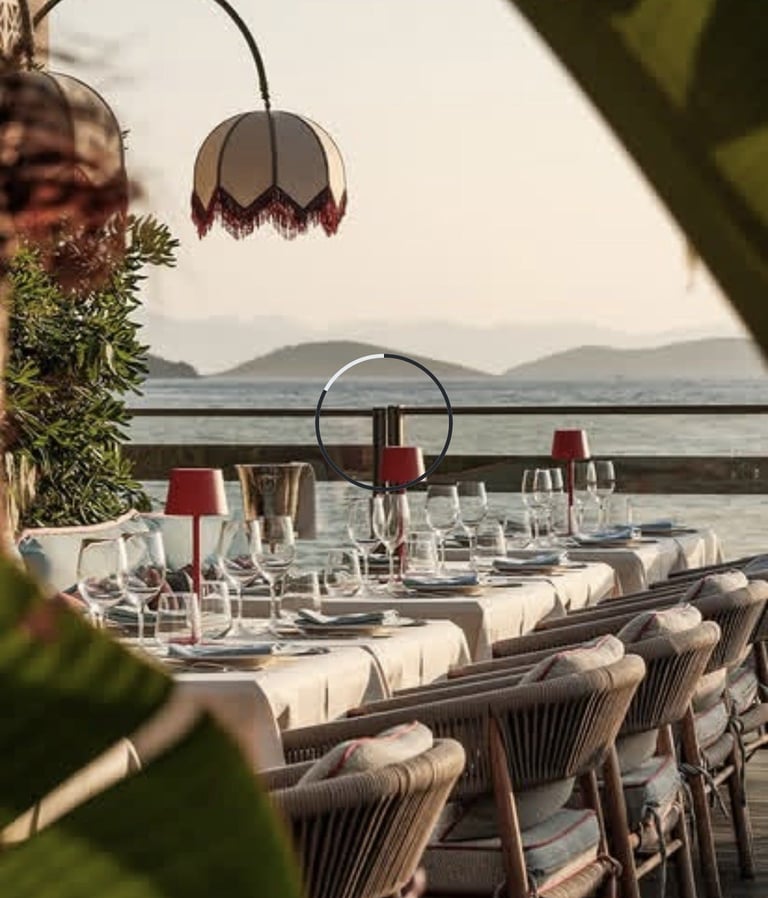 Waterfront restaurant terrace with set dining tables, pendant lamps, and mountain views across calm water