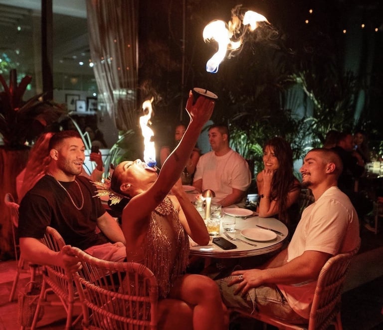Group of people dining outdoors at night with a fire performer breathing flames above their table