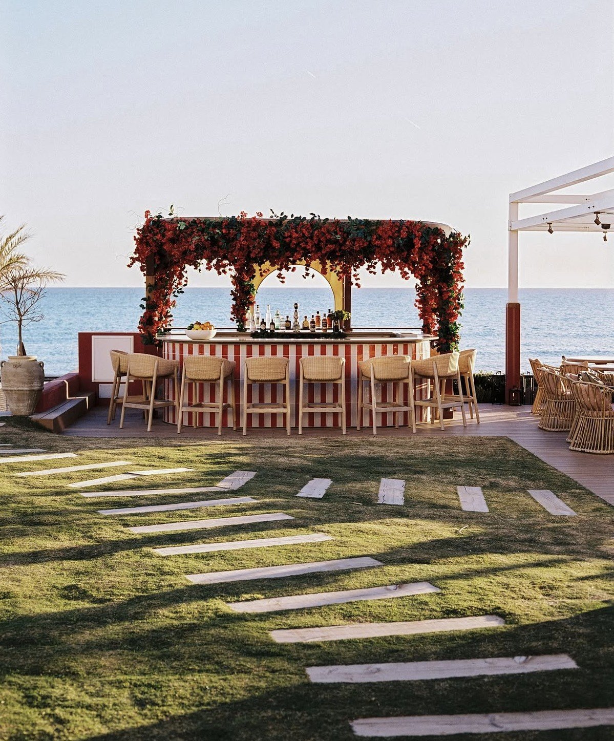 Beachfront outdoor wedding reception setup with floral-draped pergola, wooden dining table, chairs, and stone pathway on grass overlooking the sea