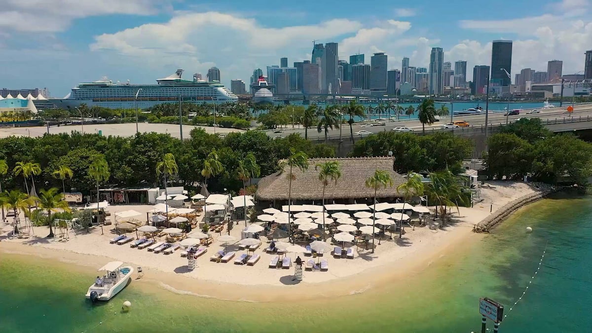 Beach with lounge chairs and umbrellas, palm trees, boat in turquoise water, city skyline in background