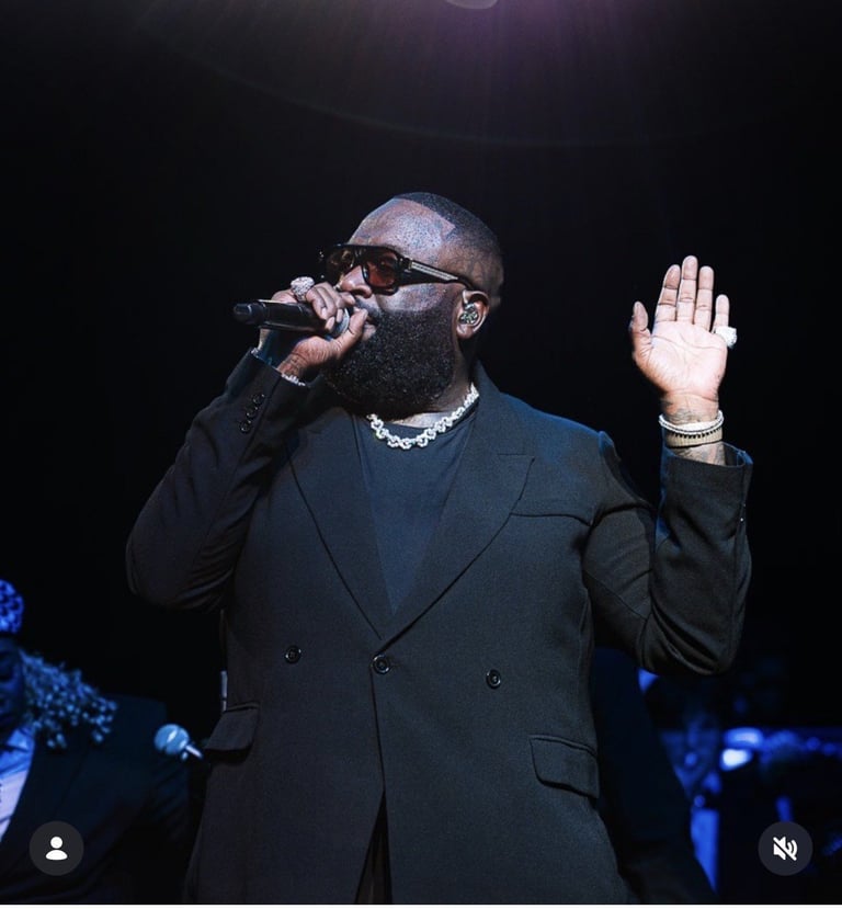 Male performer on stage singing into microphone, wearing dark suit and chain necklace, right hand raised during live concert performance