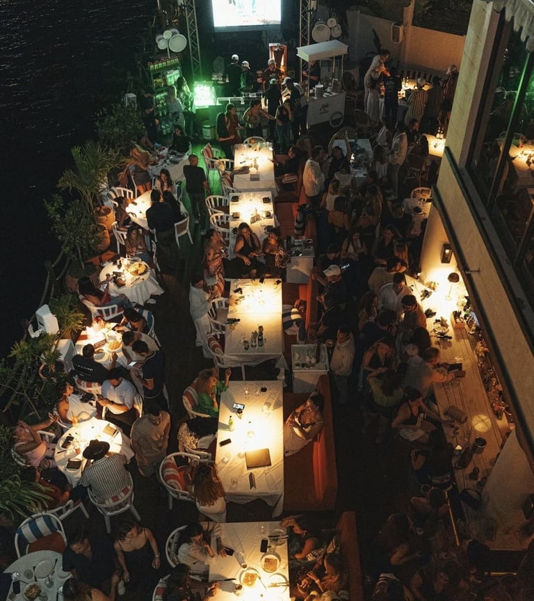 Overhead view of a lively nighttime outdoor gathering with guests dining and socializing at illuminated tables along a narrow alley