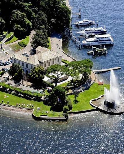 Aerial view of a lakeside villa with manicured gardens, a jet fountain, and moored boats at the dock