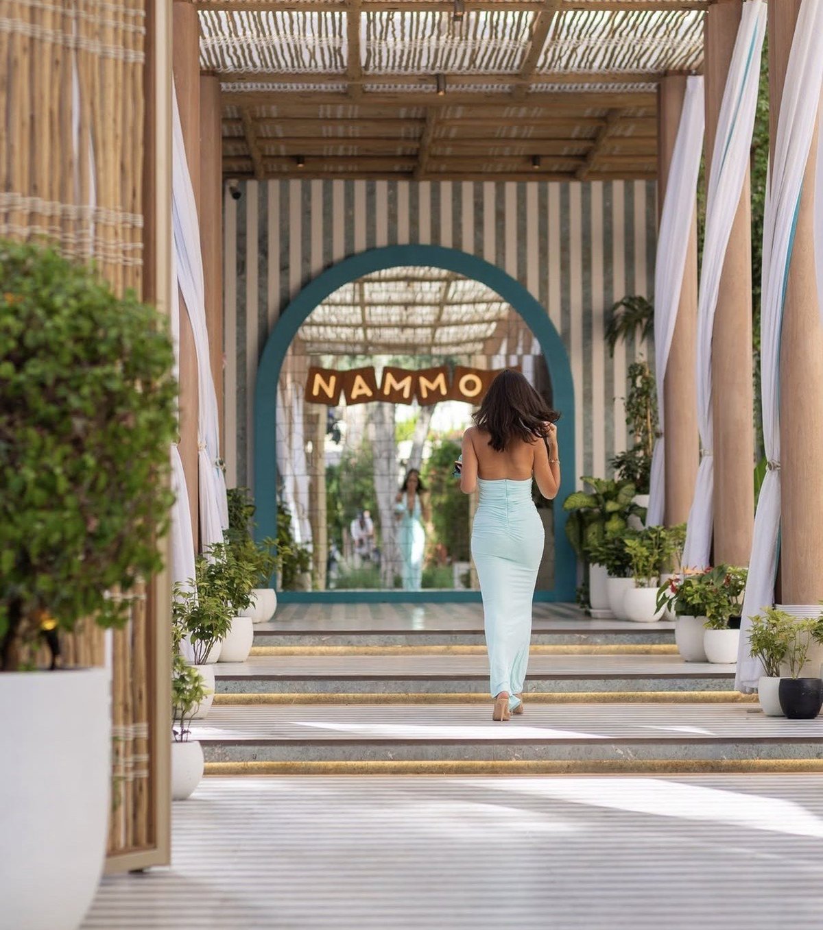 Woman in light blue dress walking through modern archway entrance with teal circular door and MAMMO signage, flanked by potted greenery and fabric drapes