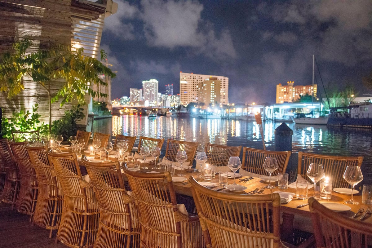 Waterfront outdoor dining area at night with wooden chairs and place settings overlooking illuminated city skyline and water