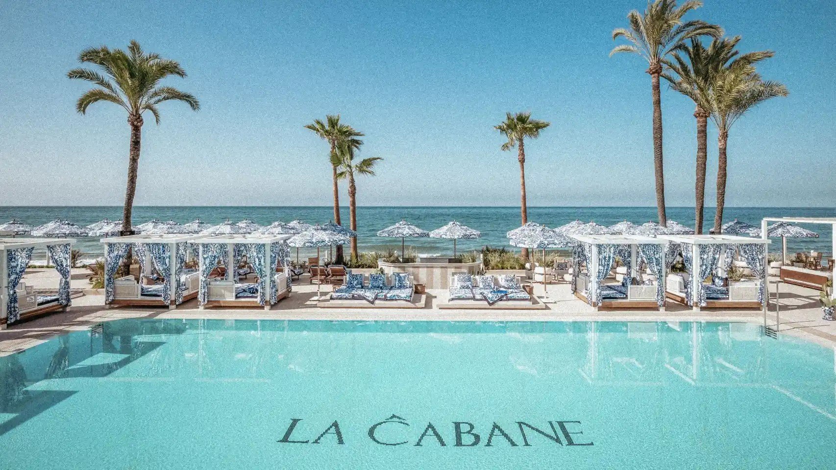 LA CABANE beach club pool with turquoise water, white loungers, palm trees, and ocean view
