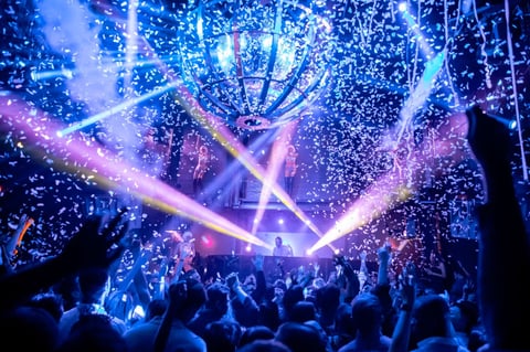 Large crowd at electronic music concert with vibrant blue and pink stage lights and sparkling confetti effects