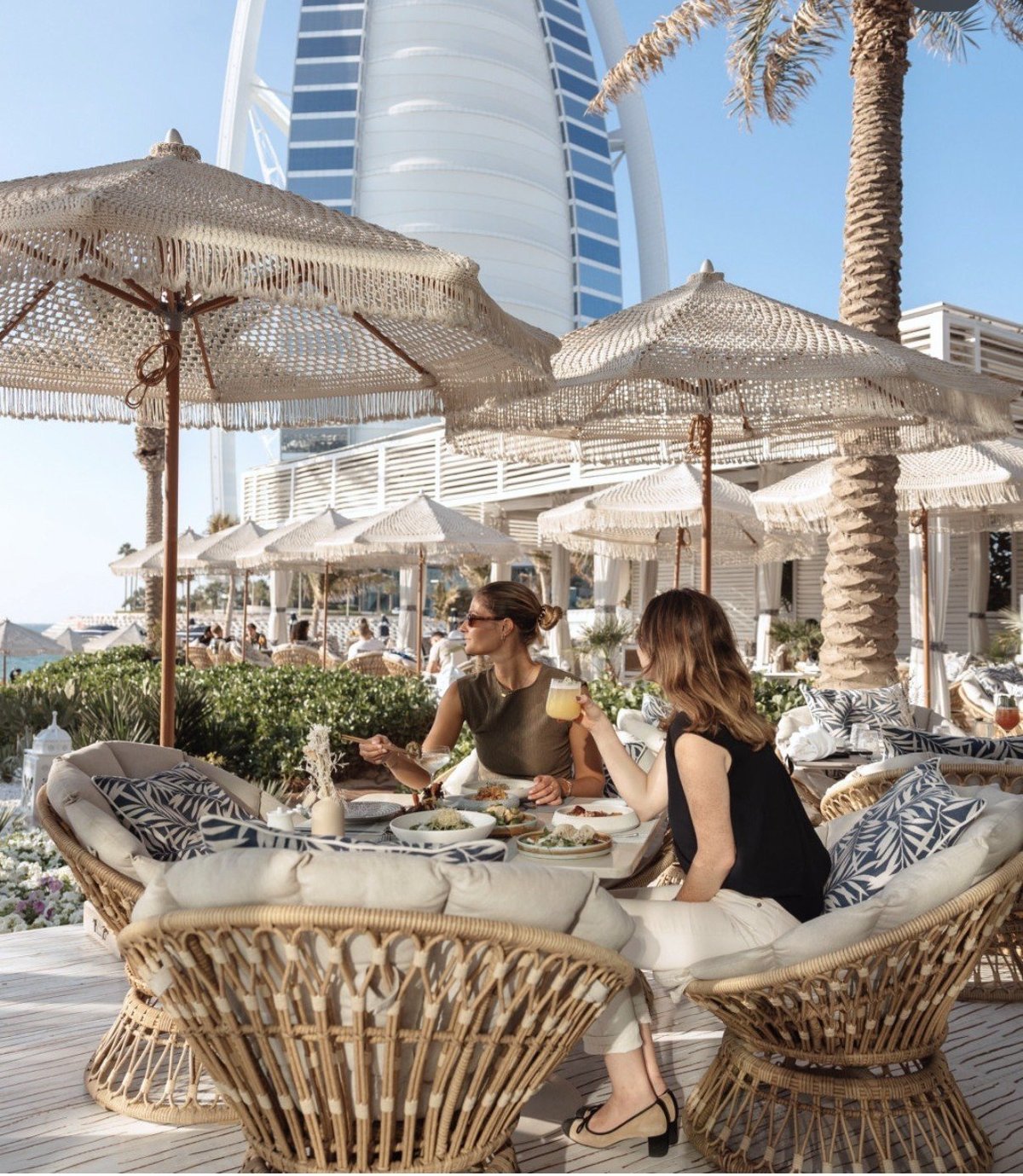 Two women dining at an outdoor beachfront restaurant with woven umbrellas, palm trees, and a luxury high-rise hotel in the background