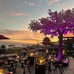 Outdoor restaurant terrace at sunset with purple-lit trees, tables with diners, and ocean view in background