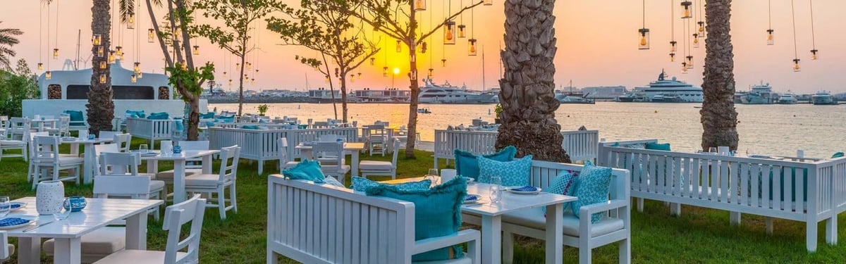Outdoor waterfront dining area at sunset with white tables, teal chairs, palm trees, and bay view