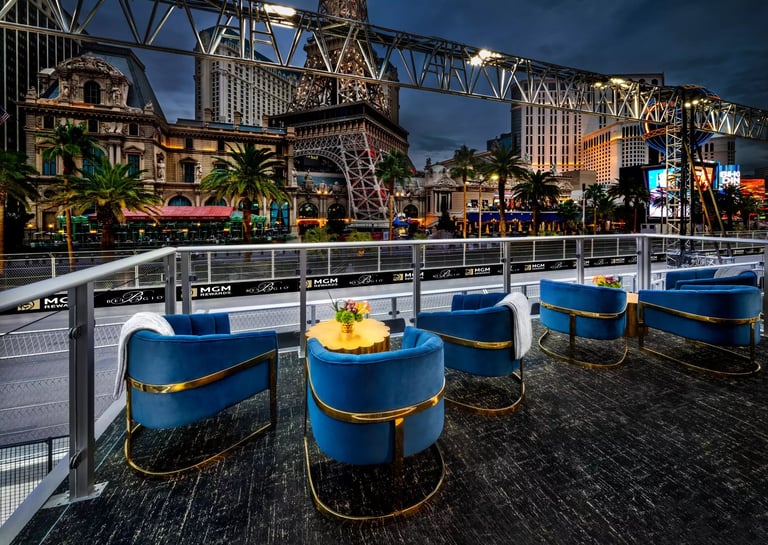 Rooftop bar with blue barrel seating and yellow tables overlooking a cityscape with ornate buildings and palm trees at dusk