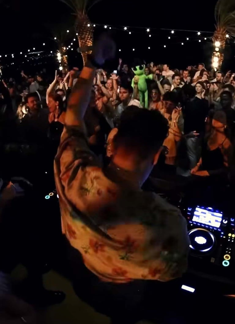 DJ performing at outdoor night event with crowd of people dancing, lit by string lights and stage lighting