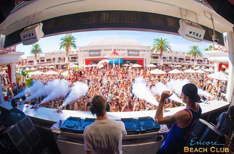 DJ booth overlooking packed beach club crowd with blue stage lights and palm trees