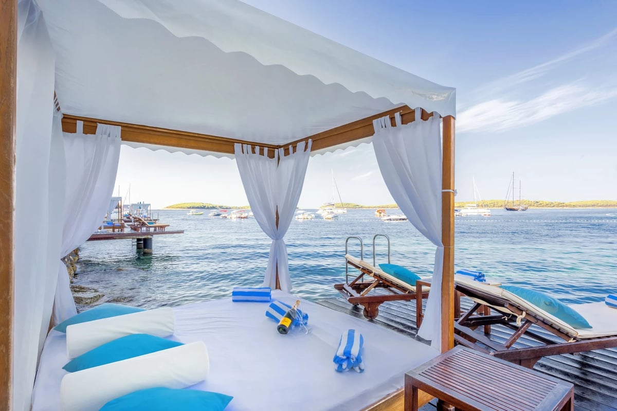 Waterfront cabana with white curtains, blue and white cushioned seating, and ocean view of moored boats