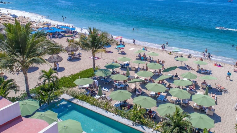 Aerial view of a tropical beach with blue umbrellas, palm trees, and a swimming pool in the foreground overlooking the turquoise ocean.