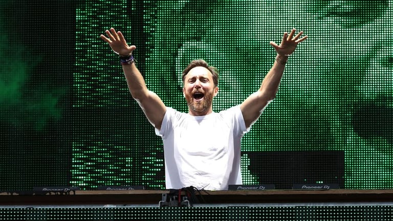 DJ with beard wearing white shirt raises arms in celebration at turntable with green digital display behind