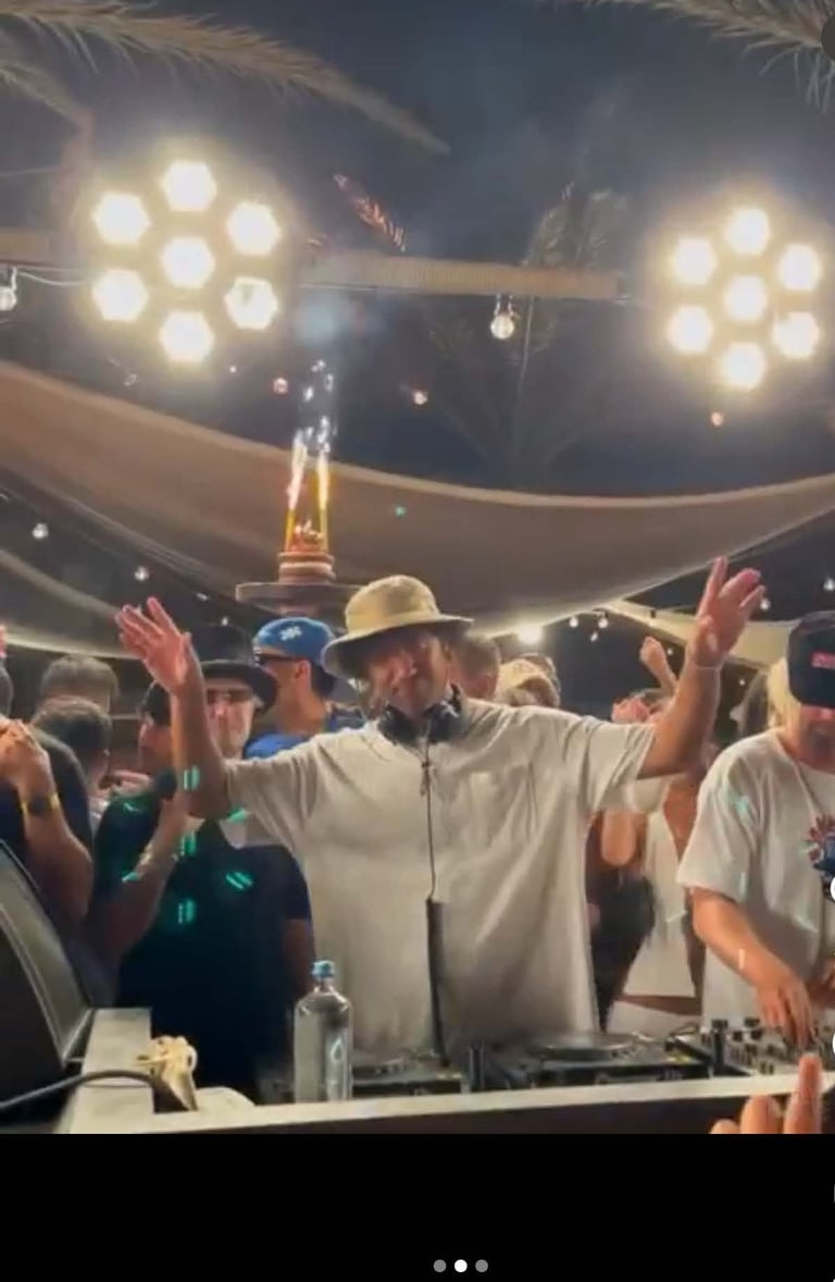 DJ in straw hat performing at nighttime outdoor event with arms raised, stage lights above, crowd around turntables