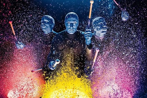 Three blue-tinted figures holding drumsticks surrounded by colorful powder particles, creating a vibrant explosion of pink, yellow, and blue colors
