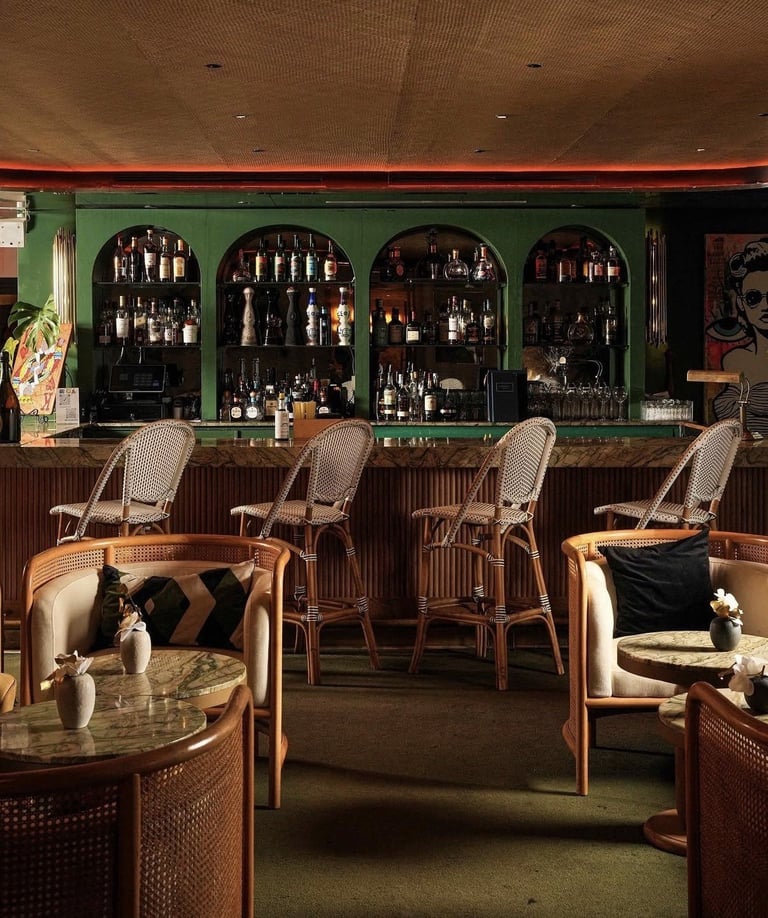 Modern bar interior with green walls, rattan chairs, and dark bottle-lined shelving behind the counter