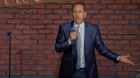 Man in dark blue suit and light blue shirt holding microphone, gesturing while performing on stage against red brick wall