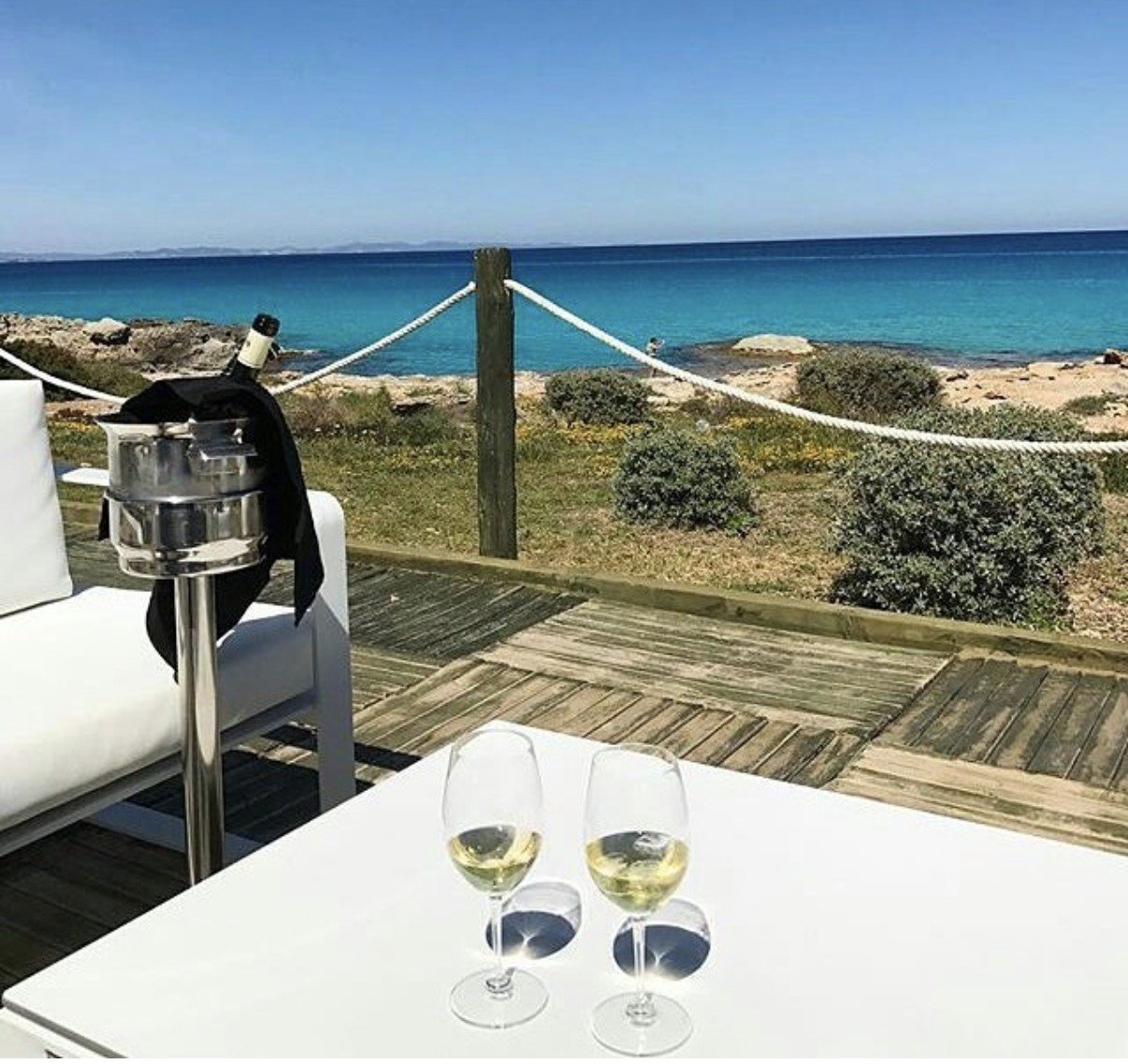 Two glasses of white wine on a table overlooking a turquoise coastal bay with wooden deck and rope railing