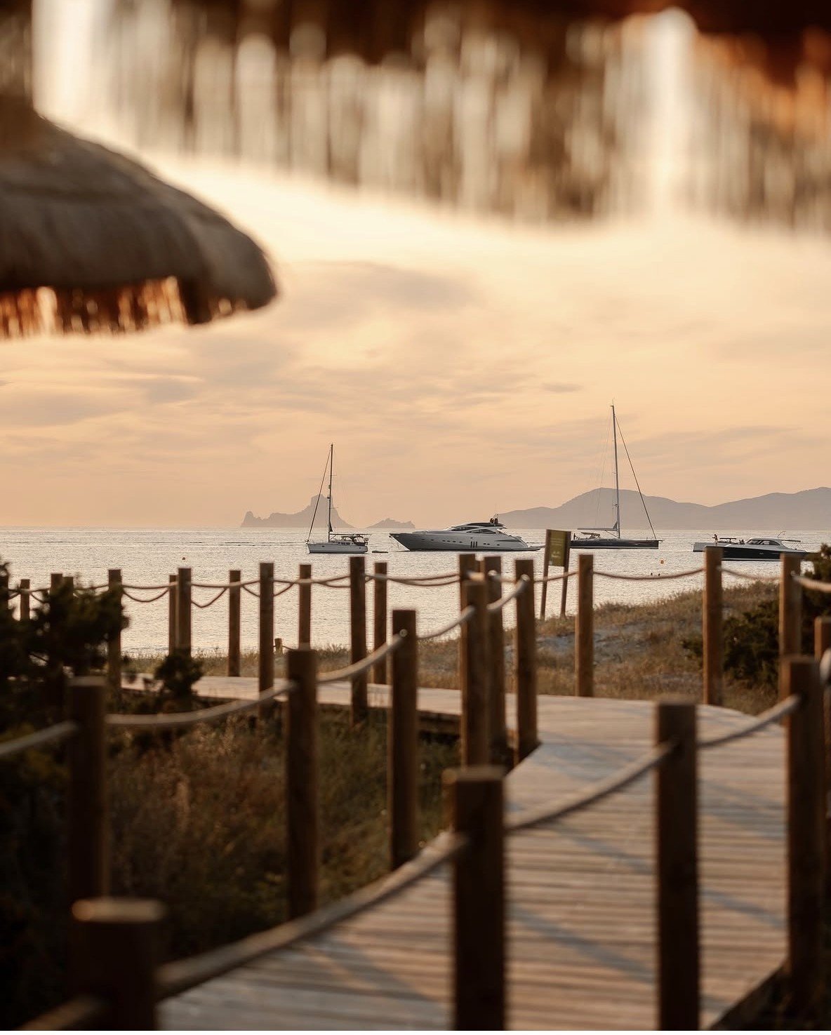 Wooden boardwalk leading to a beach with sailboats anchored in calm water and mountains in the distance