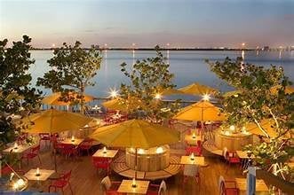 Waterfront terrace with yellow umbrellas and outdoor seating lit up at dusk overlooking a bay