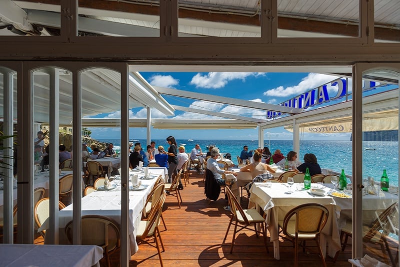Beachfront restaurant with open-air seating, diners at tables overlooking turquoise ocean water and blue sky