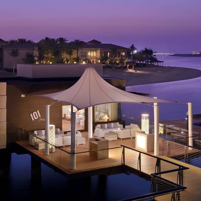 Luxury waterfront villa with tent-like roof structure at dusk, illuminated interior, moored over calm blue water with resort buildings in background