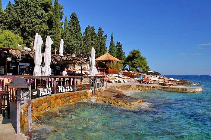 Coastal beach bar with white umbrellas, wooden deck, and rocky shoreline overlooking turquoise Adriatic Sea waters