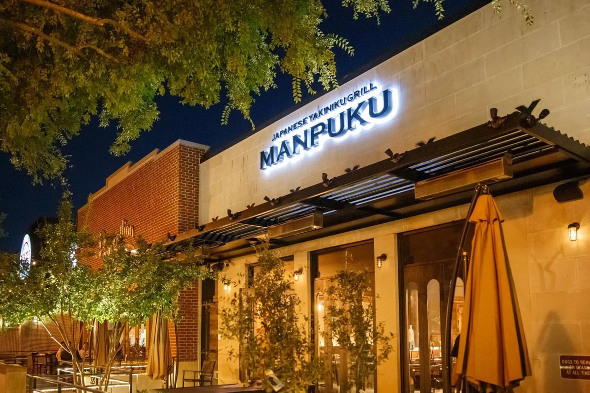 Exterior of Manfuku restaurant at night with illuminated signage, warm lighting, brick building, and trees overhead