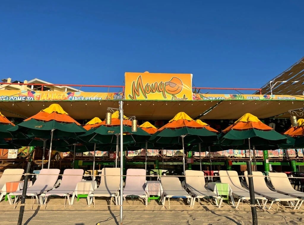 Beach bar with yellow and green umbrellas, lounge chairs, and Mango sign under clear blue sky