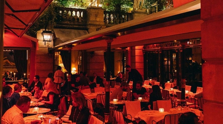 Indoor event venue with red lighting, multiple dining tables, and guests mingling in an elegant courtyard setting at night
