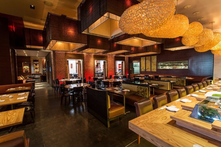 Modern restaurant interior with wooden tables, black chairs, warm ambient lighting, decorative spherical fixtures, and dark wood ceiling elements