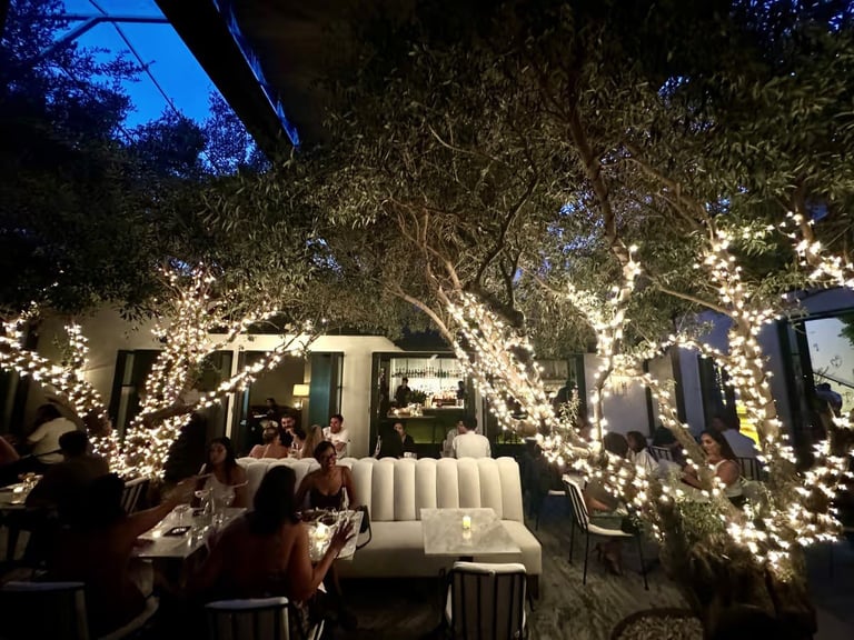 Outdoor patio at night with illuminated trees, dining tables, and elegant seating under warm string lights