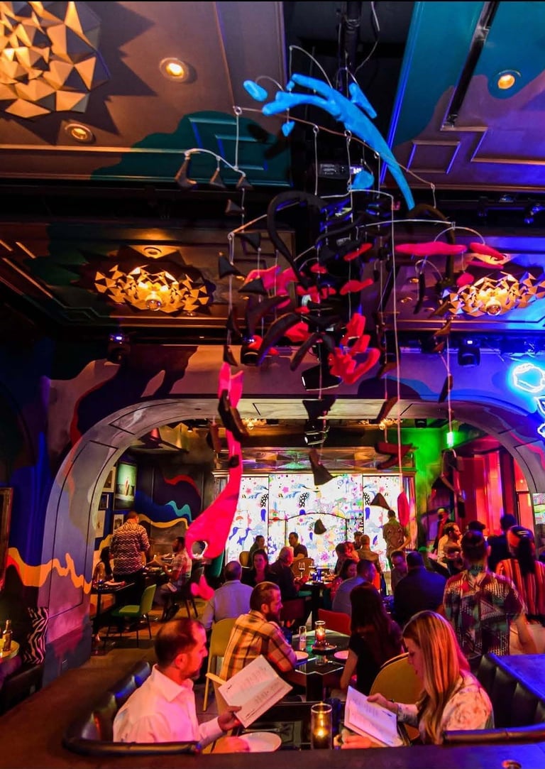 Vibrant nightclub interior with colorful neon lighting, crowds of people dancing and socializing, and artistic installations overhead.