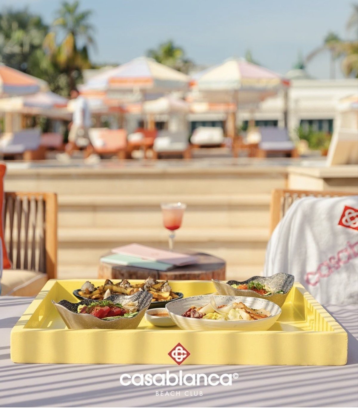 Casablanca branded yellow tray with three food compartments served outdoors on a striped table, overlooking a resort poolside