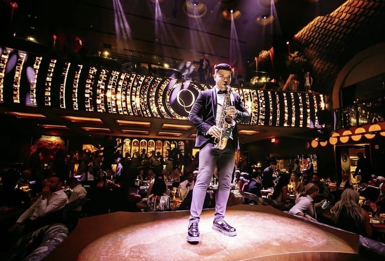 Saxophonist performing on a lit stage surrounded by seated audience members in an ornate venue with decorative lighting