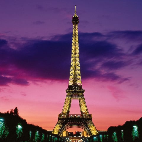 Illuminated Eiffel Tower at dusk with vibrant purple and pink sky and green-lit tree-lined avenue leading to it