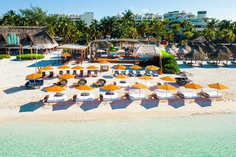Beachfront resort with rows of yellow umbrellas and loungers on sand, turquoise water, palm trees, and thatched buildings in background
