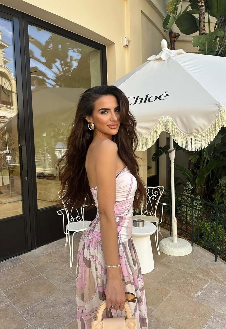 Woman with long dark hair wearing a pink and white patterned dress poses outdoors on a patio near a white umbrella and storefront