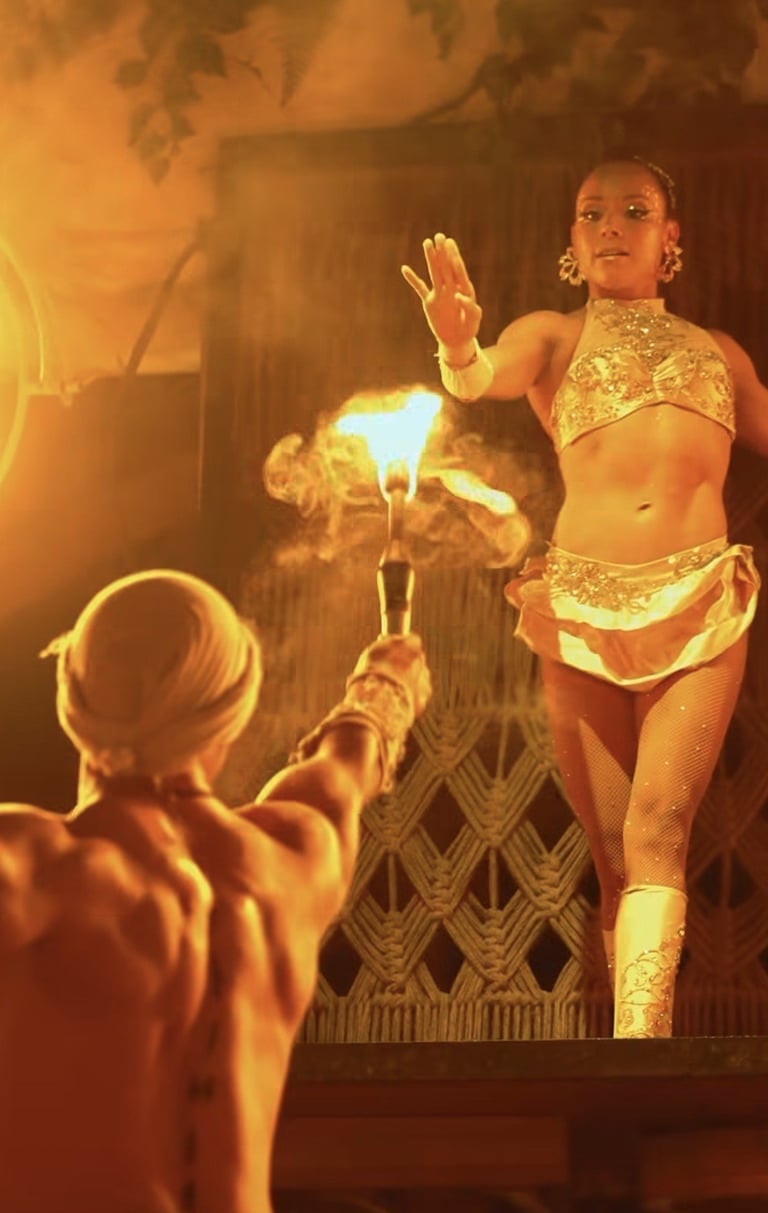 Fire performer in gold costume gestures expressively while juggling flames on stage in warm amber lighting