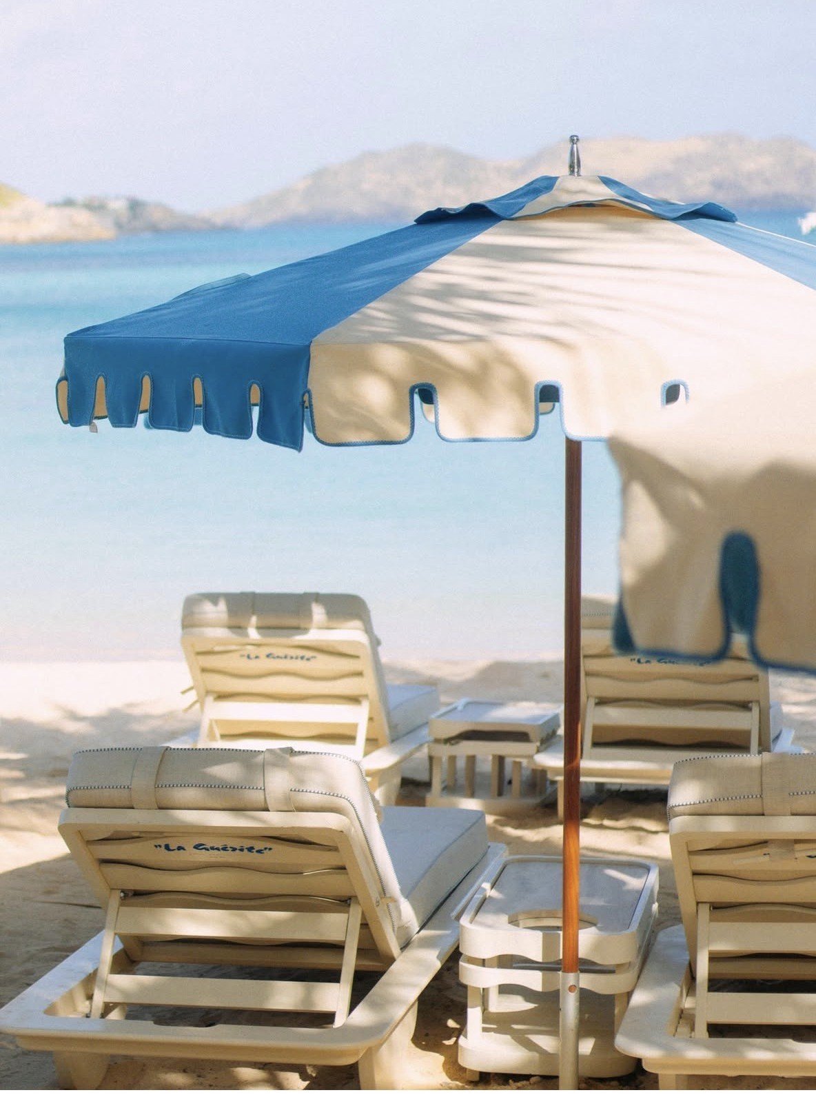 Beach scene with blue and white striped umbrella and lounge chairs overlooking turquoise ocean and distant coastline