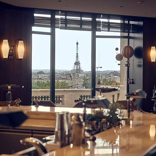 Elegant interior with Eiffel Tower view through large windows, wooden bar counter, warm lighting, and Paris cityscape